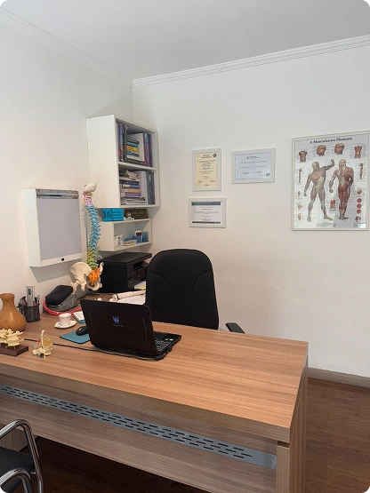 Sala de atendimento com mesa de trabalho, diplomas na parede e modelo de coluna vertebral.